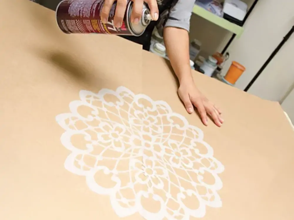 A hand spraying Sprayidea temporary adhesive onto a intricate mandala stencil placed on a large brown surface, preparing for multi-layer stenciling