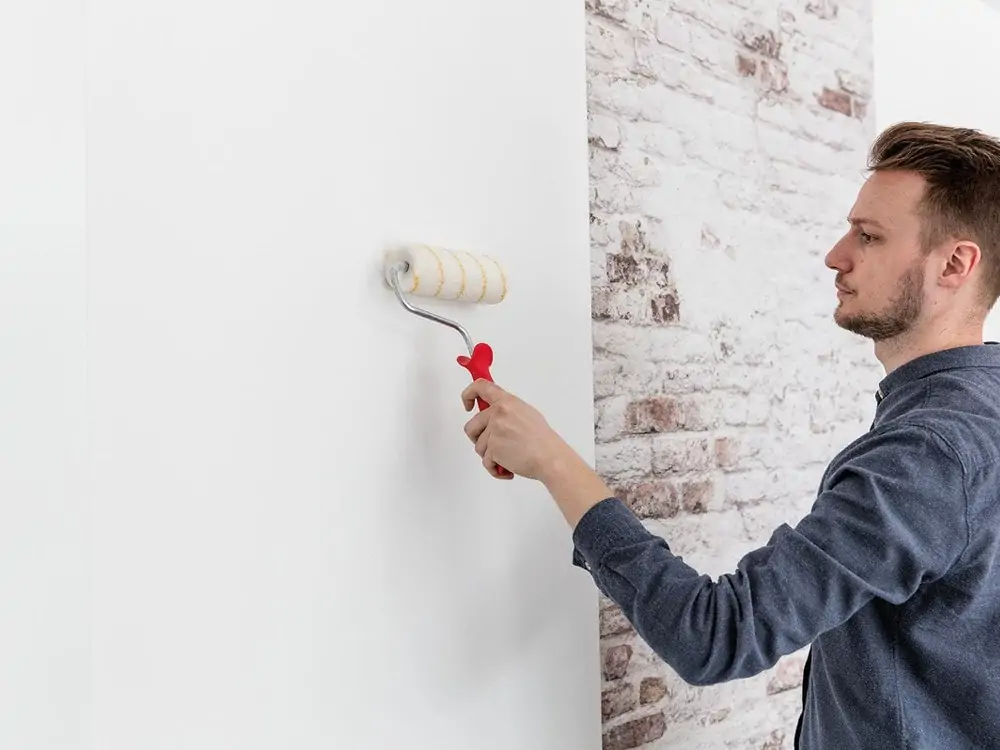 Person using a paint roller to apply adhesive or primer to a clean white wall, a crucial step to prevent wallpaper from falling off after installation.